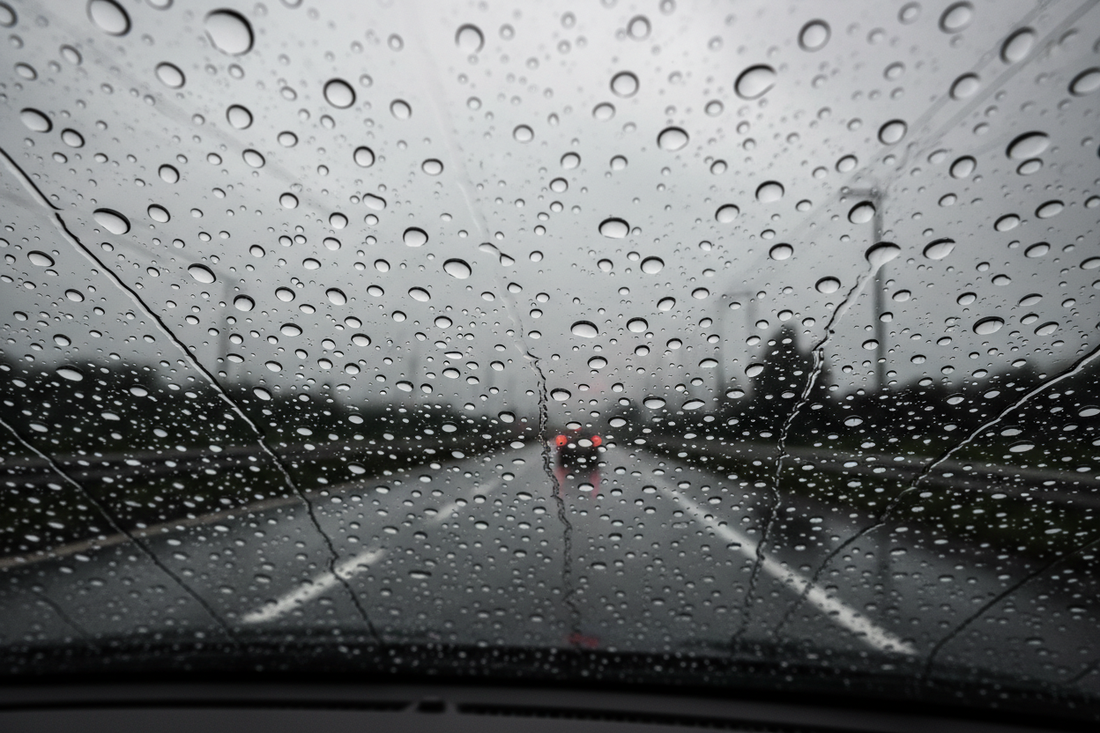 Water beading on a treated car windshield — large water droplets beading and rolling off a hydrophobic coated windshield at highway speed in rain
