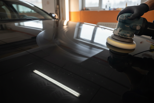 Dual-action orbital polisher being used on a dark car hood in a garage — removing swirl marks from paint with warm lighting